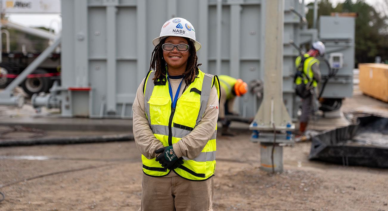 Woman wearing a bright yellow safety vest smiles while working on the construction of Harpers Switching Station