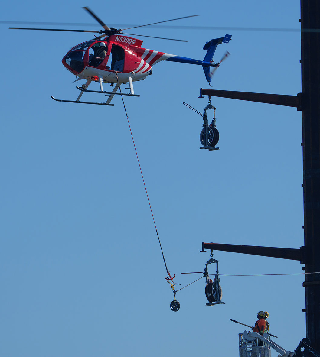 A helicopter flies above transmission towers and carries a rope and pully to assists with installing new transmission lines