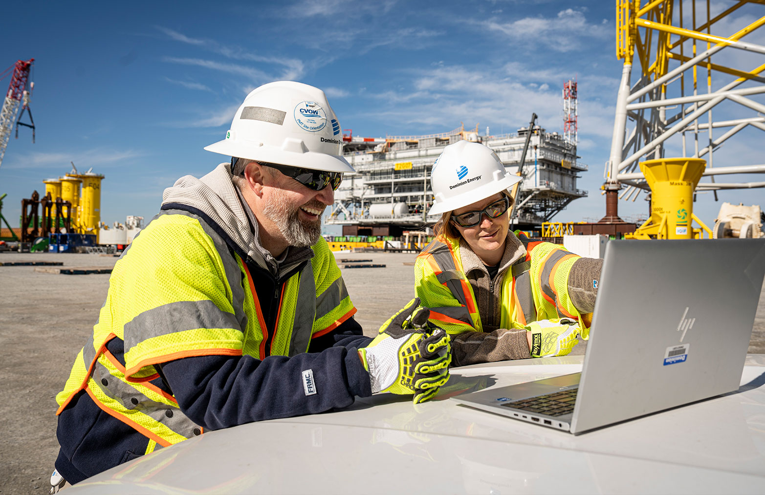 Two Dominion Energy employees in safety gear look at materials on a laptop with Portsmouth Martine Terminal in the background